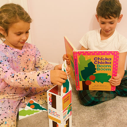 Two children sitting on the floor, one reading a book titled 'Chicka Chicka Boom Boom' and the other holding a colorful book stand.