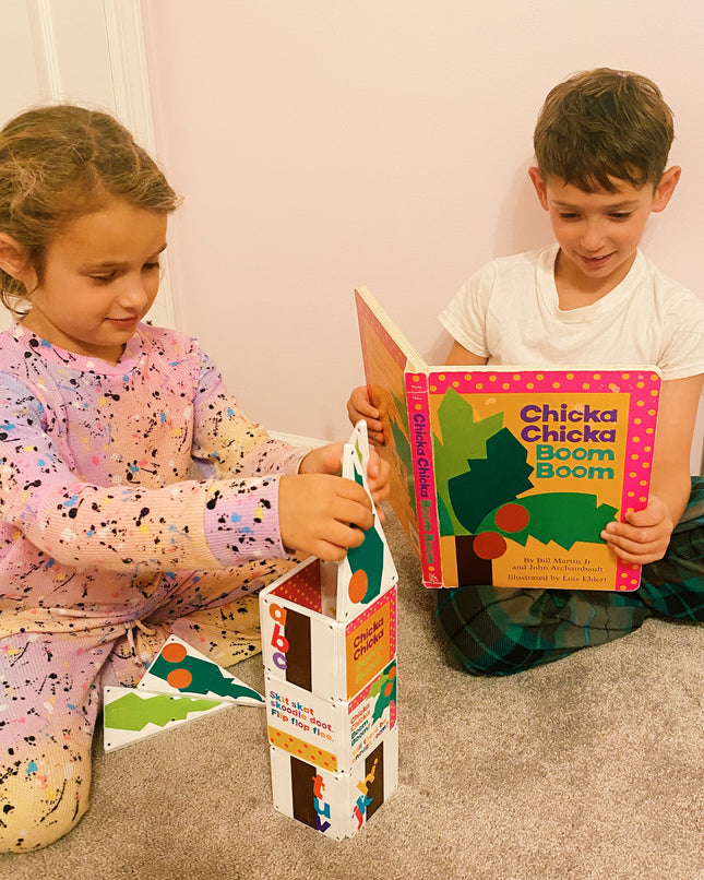 Two children sitting on the floor, one reading a book titled 'Chicka Chicka Boom Boom' and the other holding a colorful book stand.