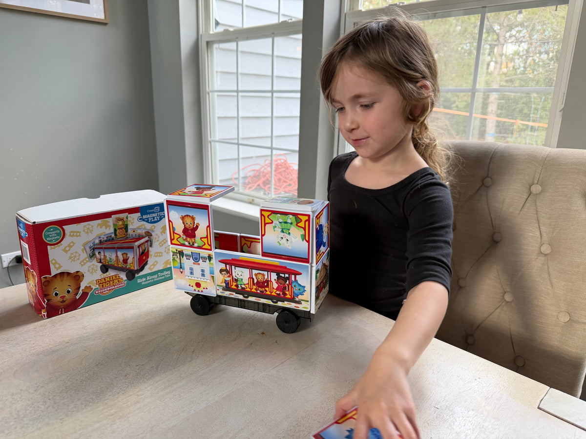 Child playing with a toy train set on a table, with toy boxes in the background.